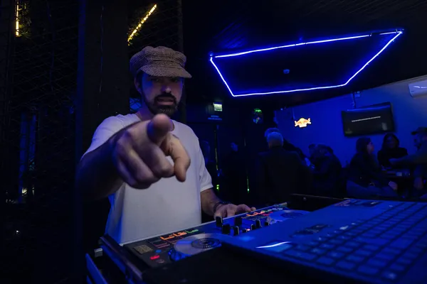 DJ de boné xadrez aponta para a câmera atrás da mesa de som, sob o neon azul