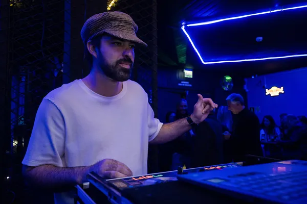DJ de boné xadrez opera a mesa de som com a mão erguida, sob o neon azul