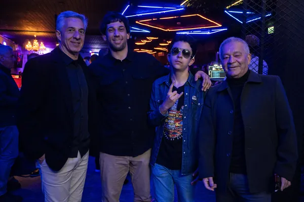 Quatro homens posam sob o neon do teto, o rapaz de óculos escuros fazendo o sinal de rock