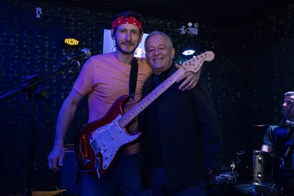 Guitarrista de bandana vermelha, com a guitarra no colo, posa abraçado a um convidado
