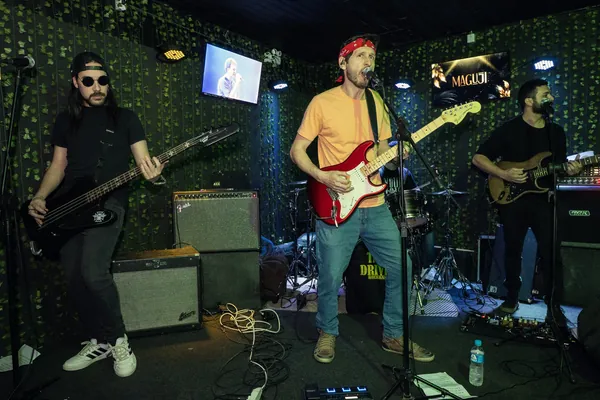 Banda no palco do Maguji, com o vocalista de bandana vermelha e camiseta laranja ao centro, entre o baixista e o guitarrista