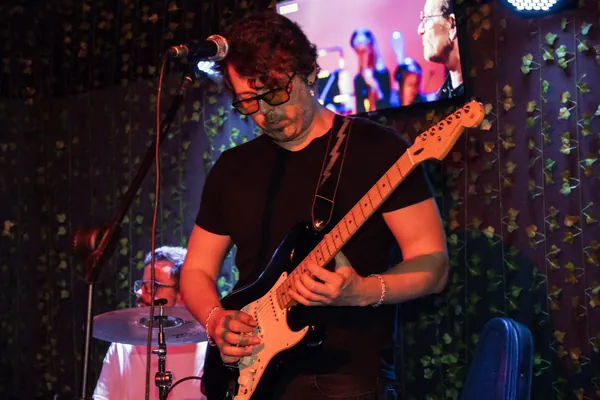 Guitarrista de óculos toca olhando para o braço da guitarra, com o baterista ao fundo