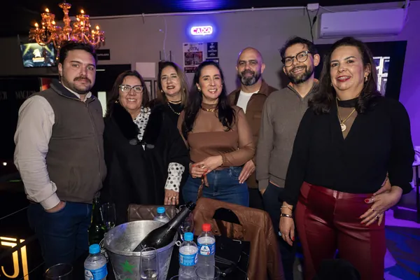 Sete convidados posam atrás da mesa com balde de gelo, sob o letreiro de neon e o lustre do salão