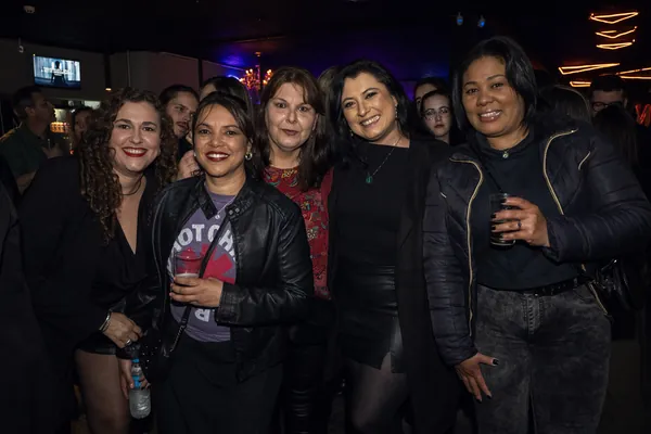 Cinco mulheres posam juntas na pista cheia, duas delas segurando copos