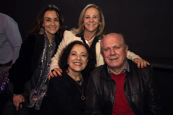 Quatro convidados posam abraçados, três mulheres e um homem de jaqueta de couro