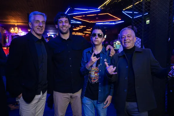 Quatro homens posam sob o neon do teto, o rapaz de óculos escuros e o senhor ao lado fazendo o sinal de rock