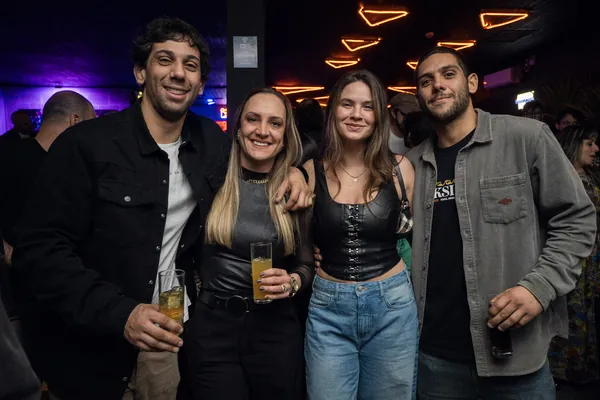 Quatro jovens posam com copos de chope, sob o neon laranja do teto