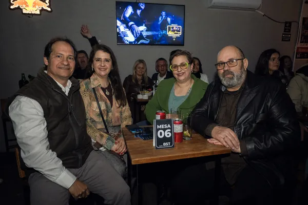 Quatro convidados sentados à mesa 06, com a TV exibindo o show ao fundo