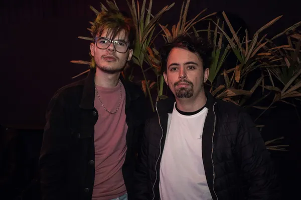 Dois jovens posam lado a lado diante da folhagem da parede