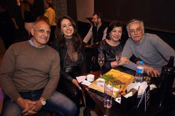 Quatro convidados sentados à mesa, com taças de espumante e petiscos servidos